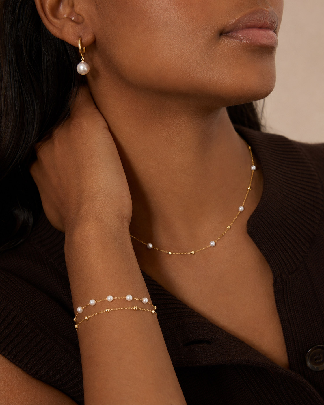 Close crop of a model wearing a drop earring, a beaded necklace, and layered bracelets with a dark brown cardigan.