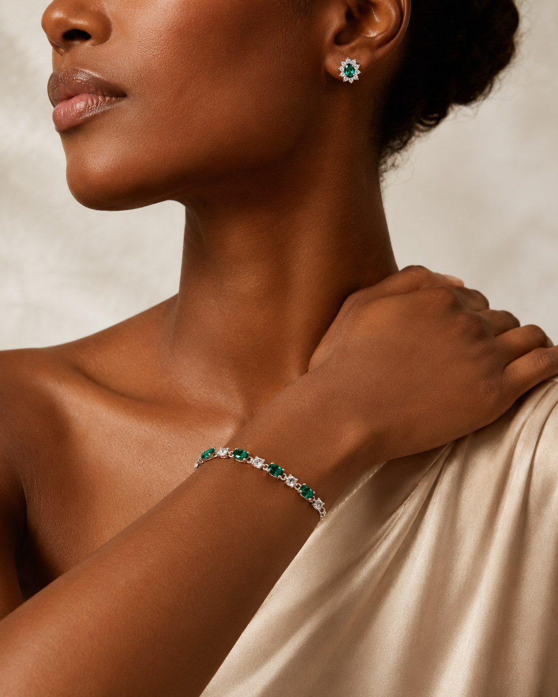 Close crop of a model wearing a green-stone earring and matching bracelet with a champagne satin one-shoulder dress.